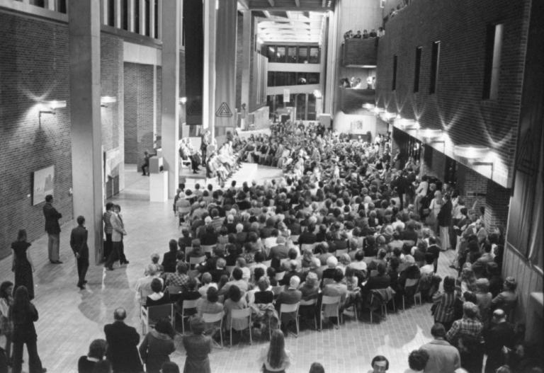 Black-and-white photo of a large audience seated in AUArts’ Main Mall (then ACA) during a formal event, with speakers on stage and people standing along the sides.