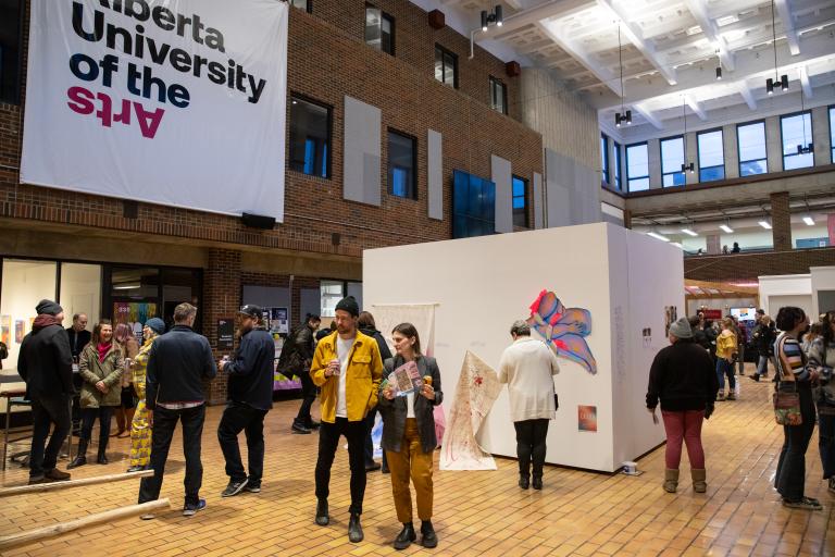 Visitors gather in AUArts’ Main Mall during the Gallery Crawl, viewing student artworks displayed on white walls beneath a large Alberta University of the Arts banner.