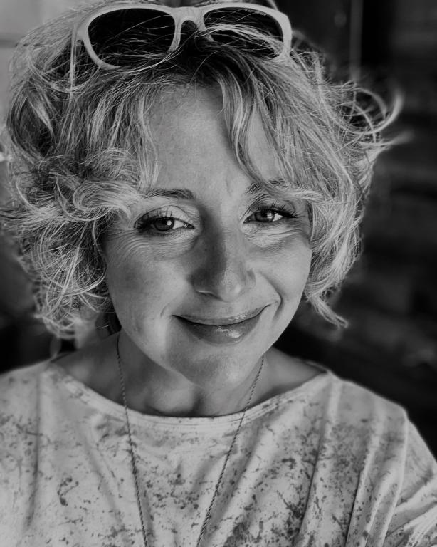 A black and white photo of Jenifer Darbellay. She has chin-length curly blonde hair and is wearing sunglasses on her head. The photo appears to be a selfie as Jenifer's gaze is not directly at the viewer. She is smiling and looks at ease.