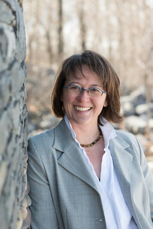 A smiling woman with short brown hair and blue-framed glasses leans against a stone wall outdoors. She wears a light grey blazer over a white blouse and a beaded necklace. The background is softly blurred with trees and rocks in natural light.
